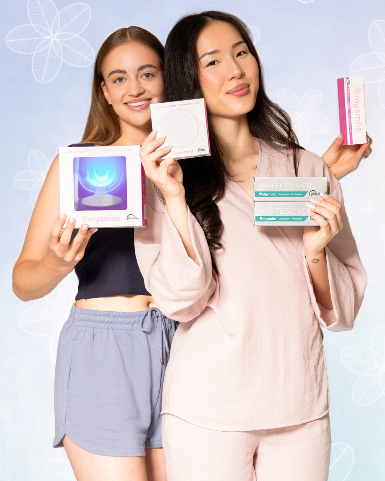 Zwei lächelnde Frauen präsentieren OnlySmile Zahnaufhellungsprodukte, darunter ein LED-Whitening-Kit, Whitening-Gel, Whitening-Pulver und WhiteSilver Zahnpasta, vor hellem Hintergrund.