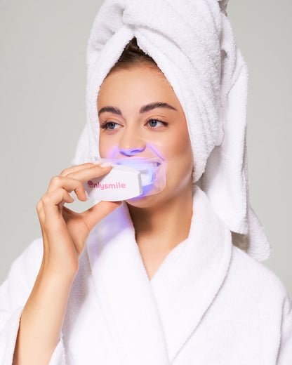 Frau im Bademantel mit Handtuch auf dem Kopf verwendet ein OnlySmile LED-Zahnaufhellungsgerät, das während der Anwendung blau leuchtet.